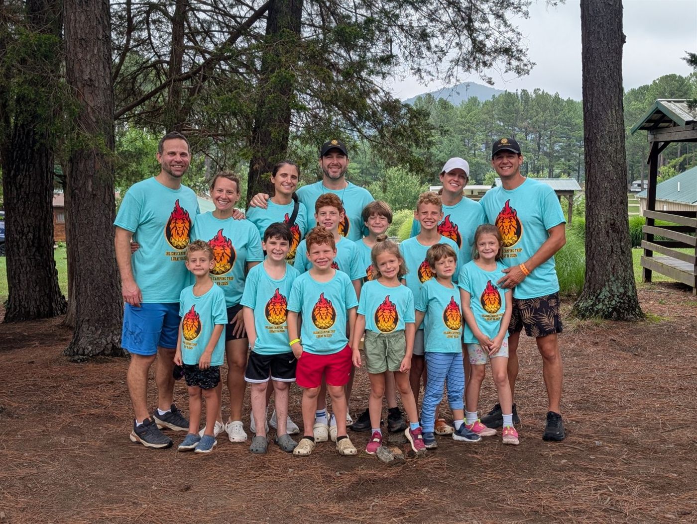Six adults and eight children in matching turquoise 18Lions camping trip shirts, posed among pine trees with a mountain ridge behind them.