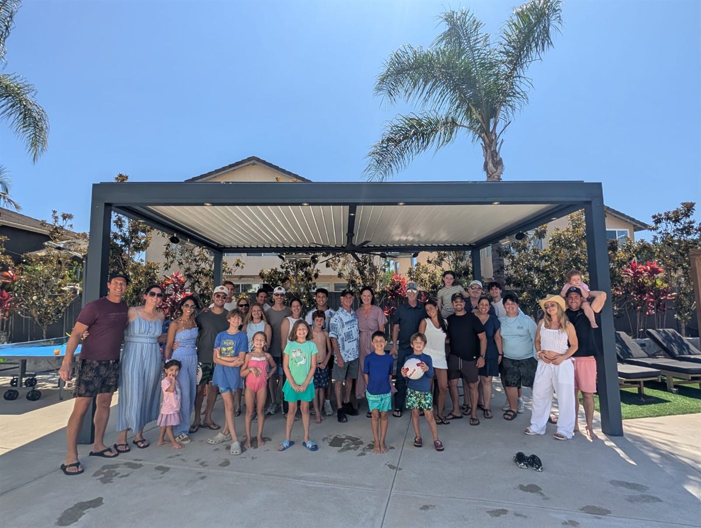 A large group of roughly thirty members, spouses, and children gathered under a pergola beside a backyard pool.